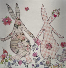 Textile bunny art showing two rabbits holding hands with soft floral accents in a gentle, romantic composition
