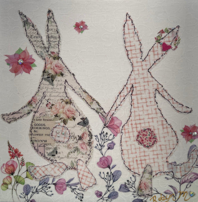 Textile bunny art showing two rabbits holding hands with soft floral accents in a gentle, romantic composition