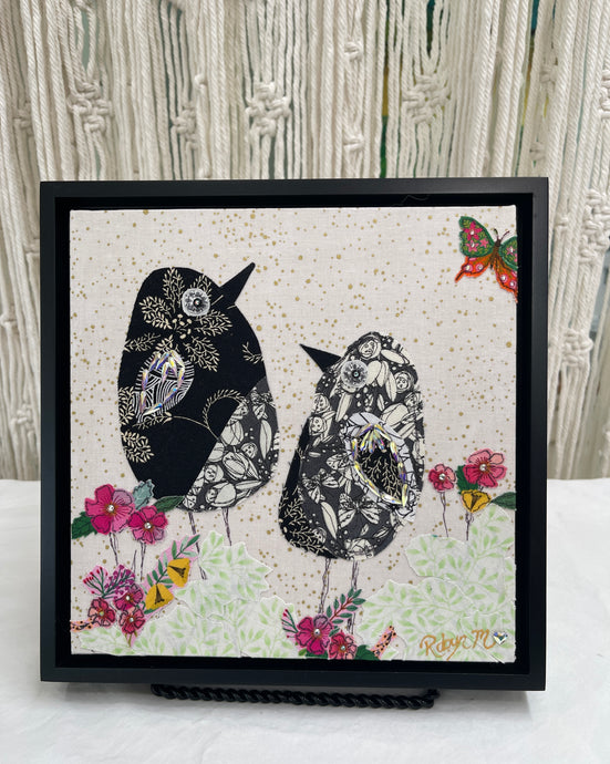 Textile bird art showing two black and white patterned birds surrounded by soft florals in a calm, minimalist composition