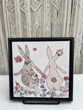 Textile bunny art showing two rabbits holding hands with soft floral accents in a gentle, romantic composition