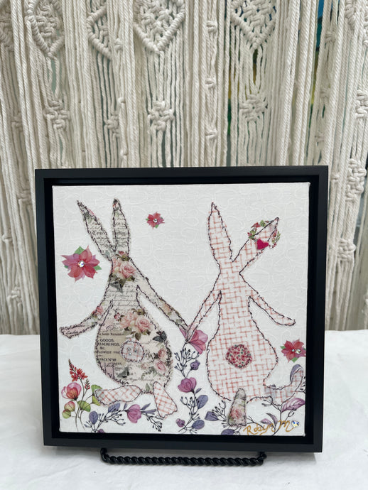 Textile bunny art showing two rabbits holding hands with soft floral accents in a gentle, romantic composition