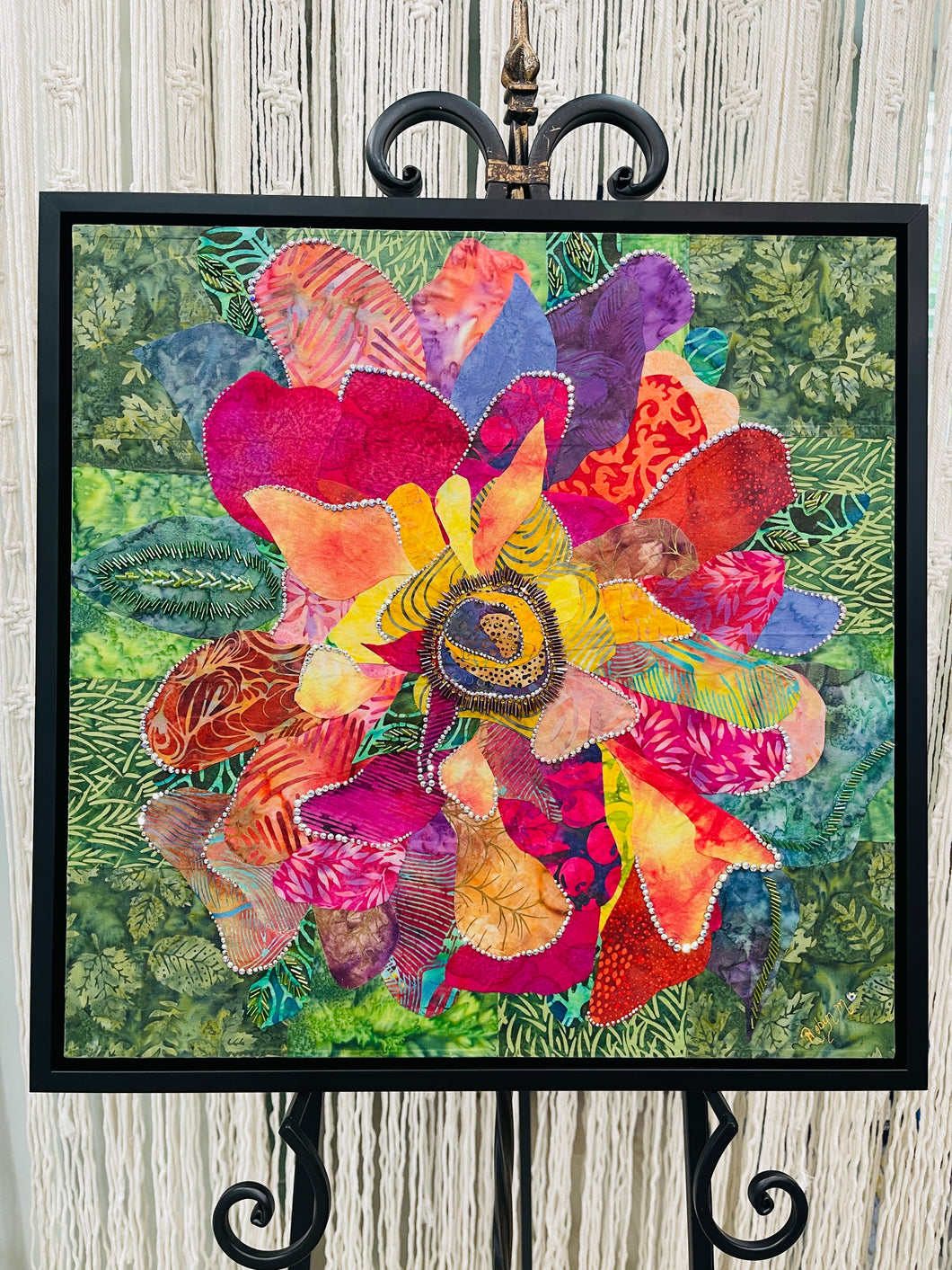 Original mixed media botanical artwork 20x20 inches, oversized flower constructed from layered batik fabrics in crimson magenta orange gold and violet, crystal outlined petals, Czech glass bead center detail with Swarovski Crystal lined petals, lush green foliage background, on birch panel, part of the Floral and Botanical Collection by Robyn Moore Arts