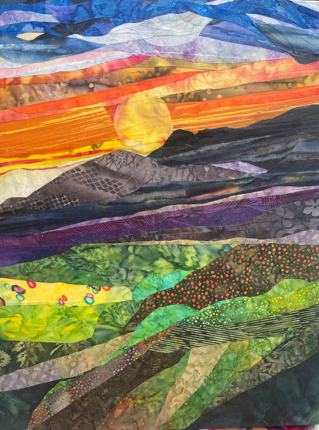 Original all fabric landscape collage artwork 20x16 inches, mountain sunset scene with golden sun descending behind dark mountain silhouettes, fiery orange and red sky, deep cobalt clouds, sweeping landscape layers in violet green and yellow batik fabric, on birch panel, part of the Where Color Takes Root Collection by Robyn Moore Arts
